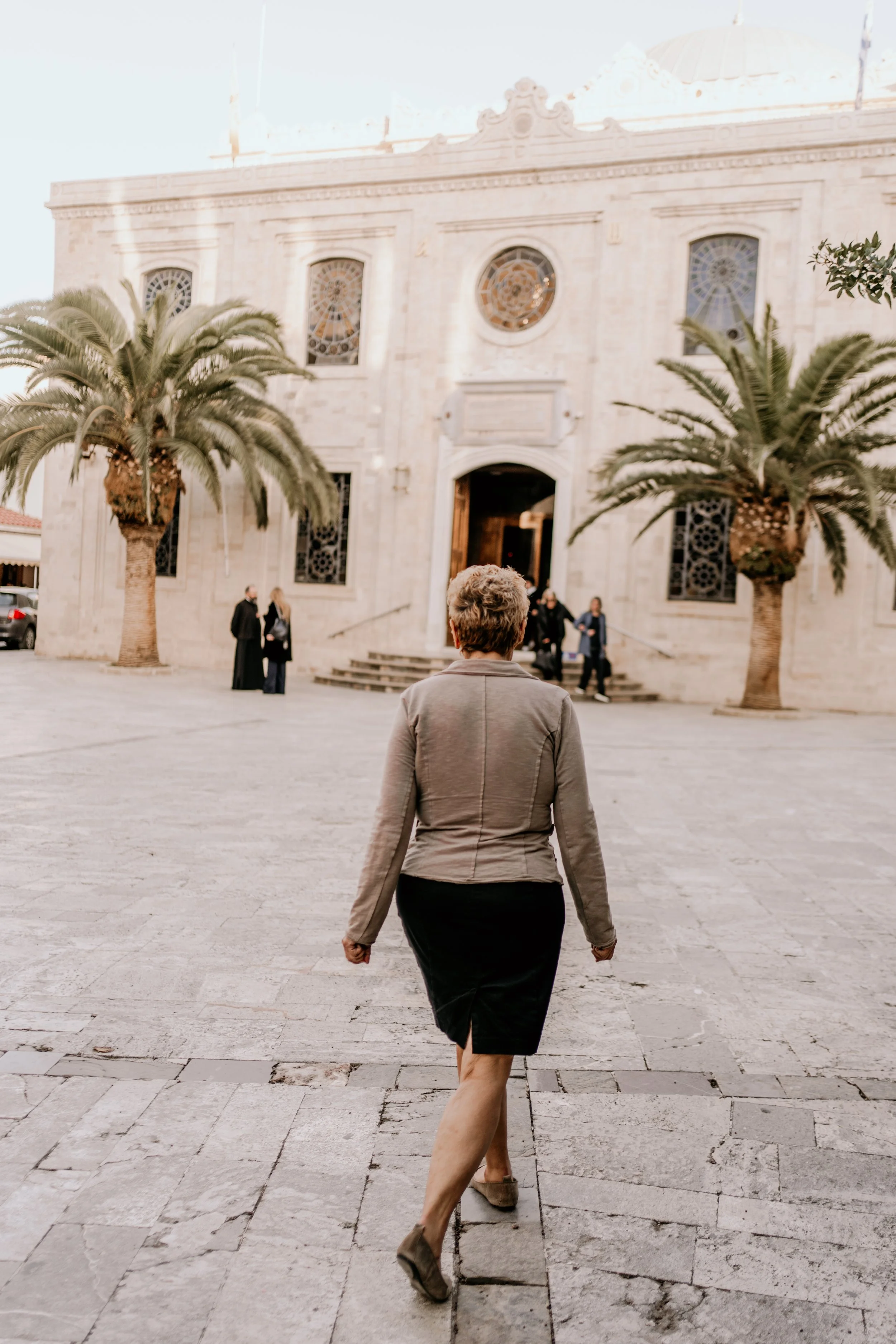 A woman with curly short hair wearing a gray blazer and black skirt walking towards a historic church with a stone facade, featuring arched windows, circular stained glass windows, and two palm trees on either side of the entrance, in a plaza with other people.