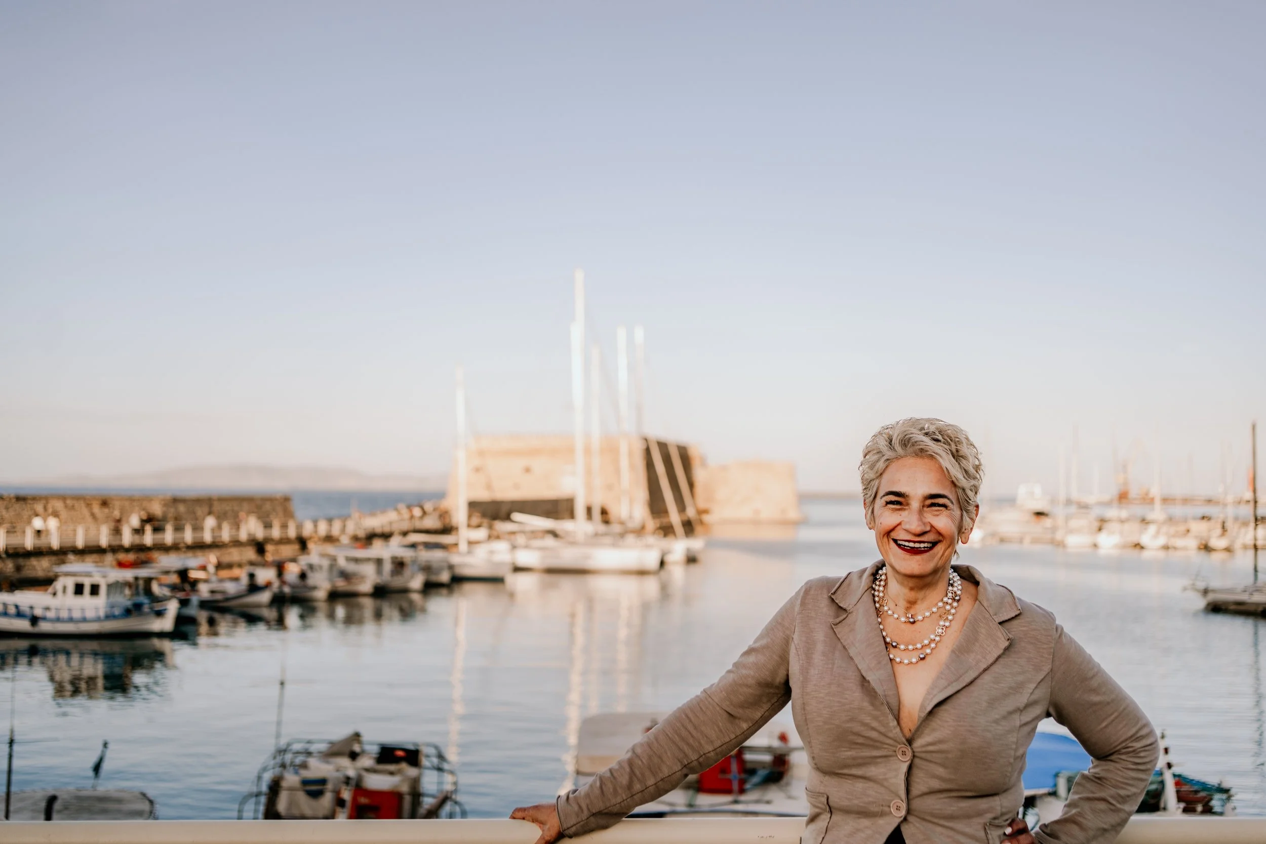 A smiling woman with short gray hair and pearl necklace standing near a marina with boats and a dock, against a backdrop of a calm body of water and a clear sky.