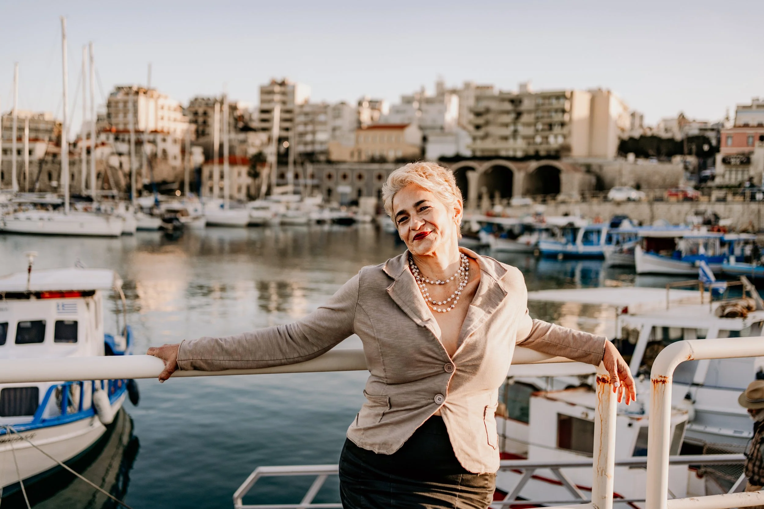 A woman with short blonde hair, wearing a beige blazer and pearl necklace, standing on a boat dock with boats and high-rise buildings in the background.
