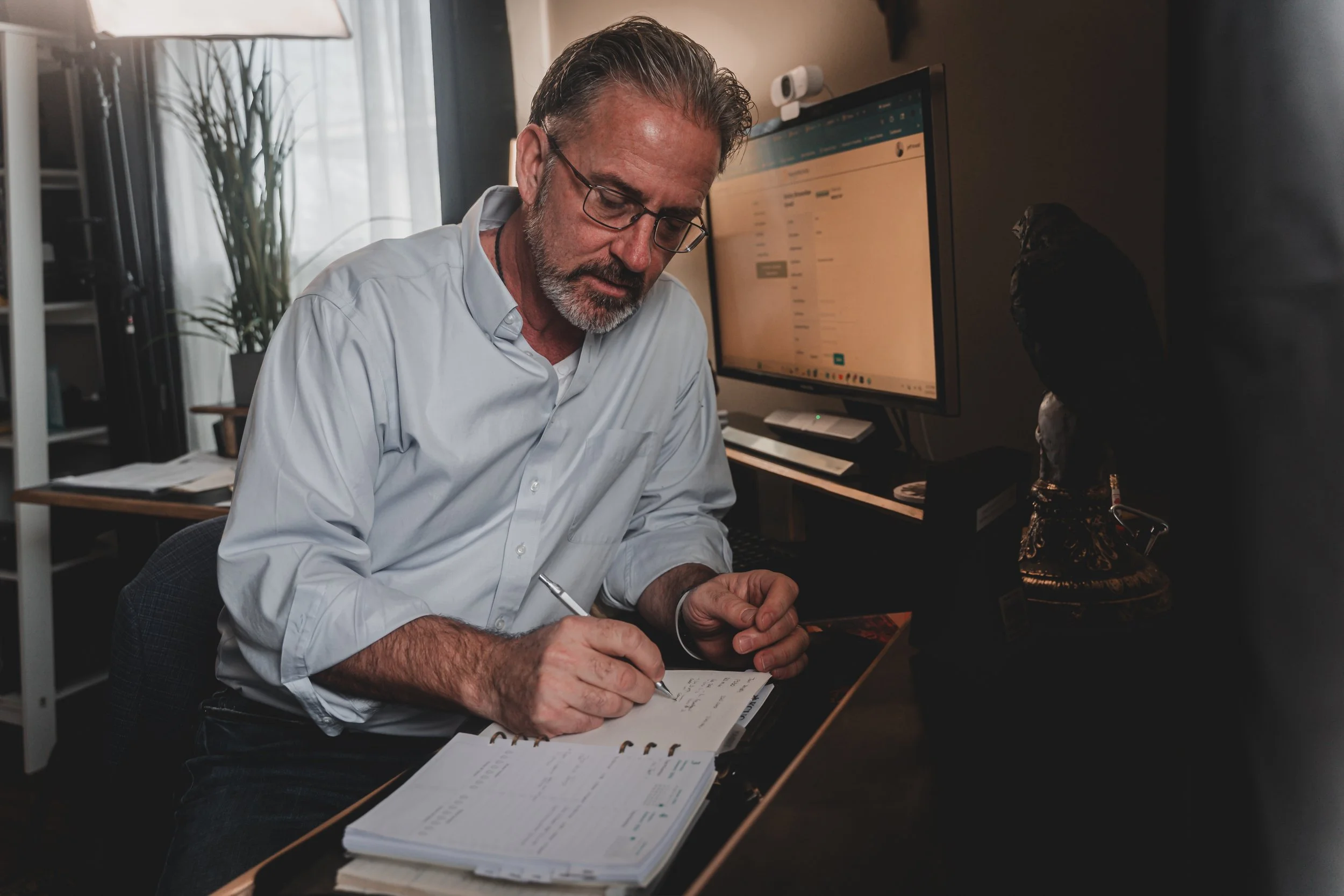 A man with glasses and a beard wearing a light blue shirt sitting at a desk, writing in a planner with a notebook open in front of him, in an office with a computer and a large monitor in the background.