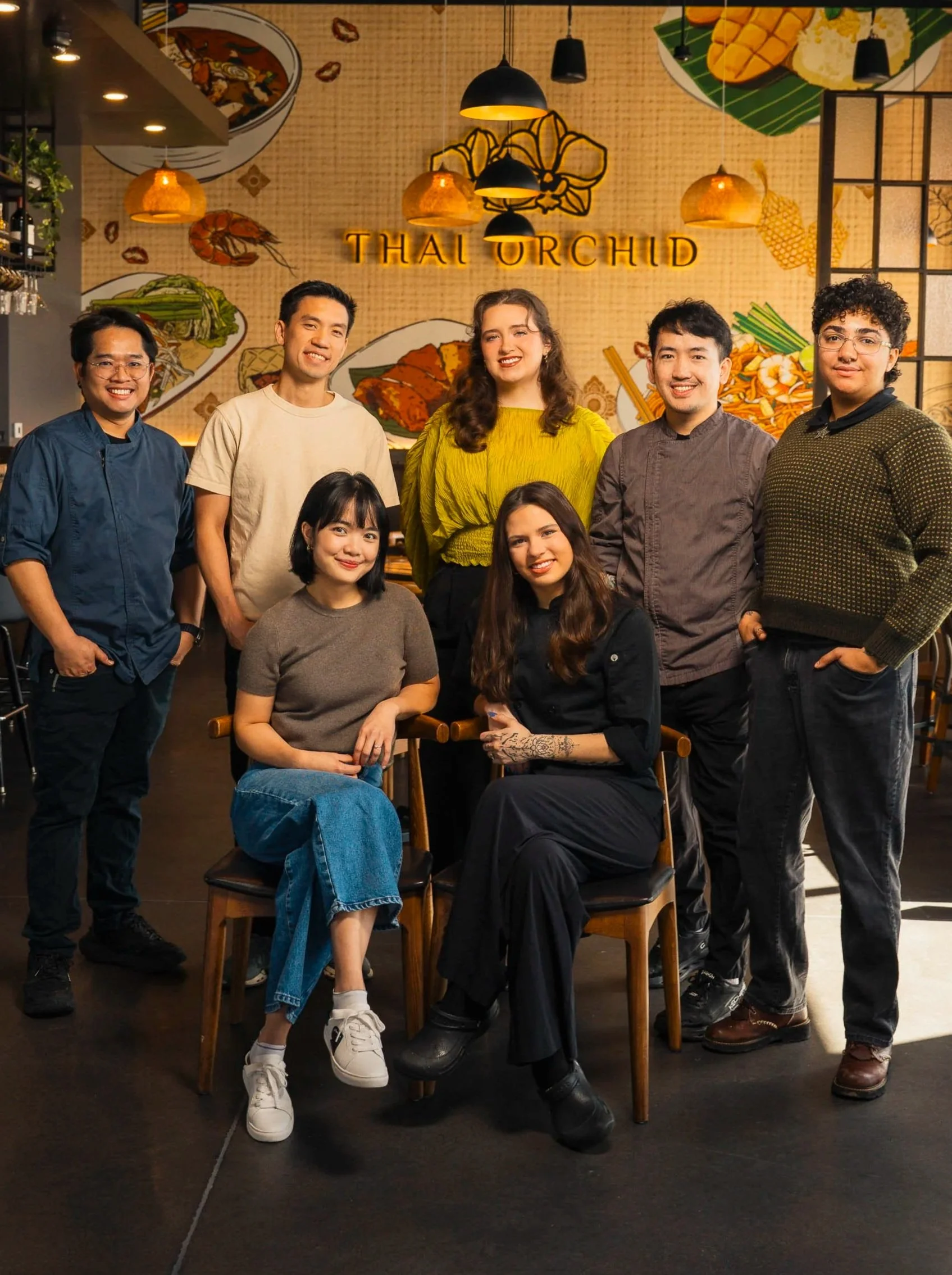 A group of eight diverse young adults in casual and semi-formal attire posing inside a restaurant named Thai Orchid. The background features Thai-themed decor and a illuminated sign.