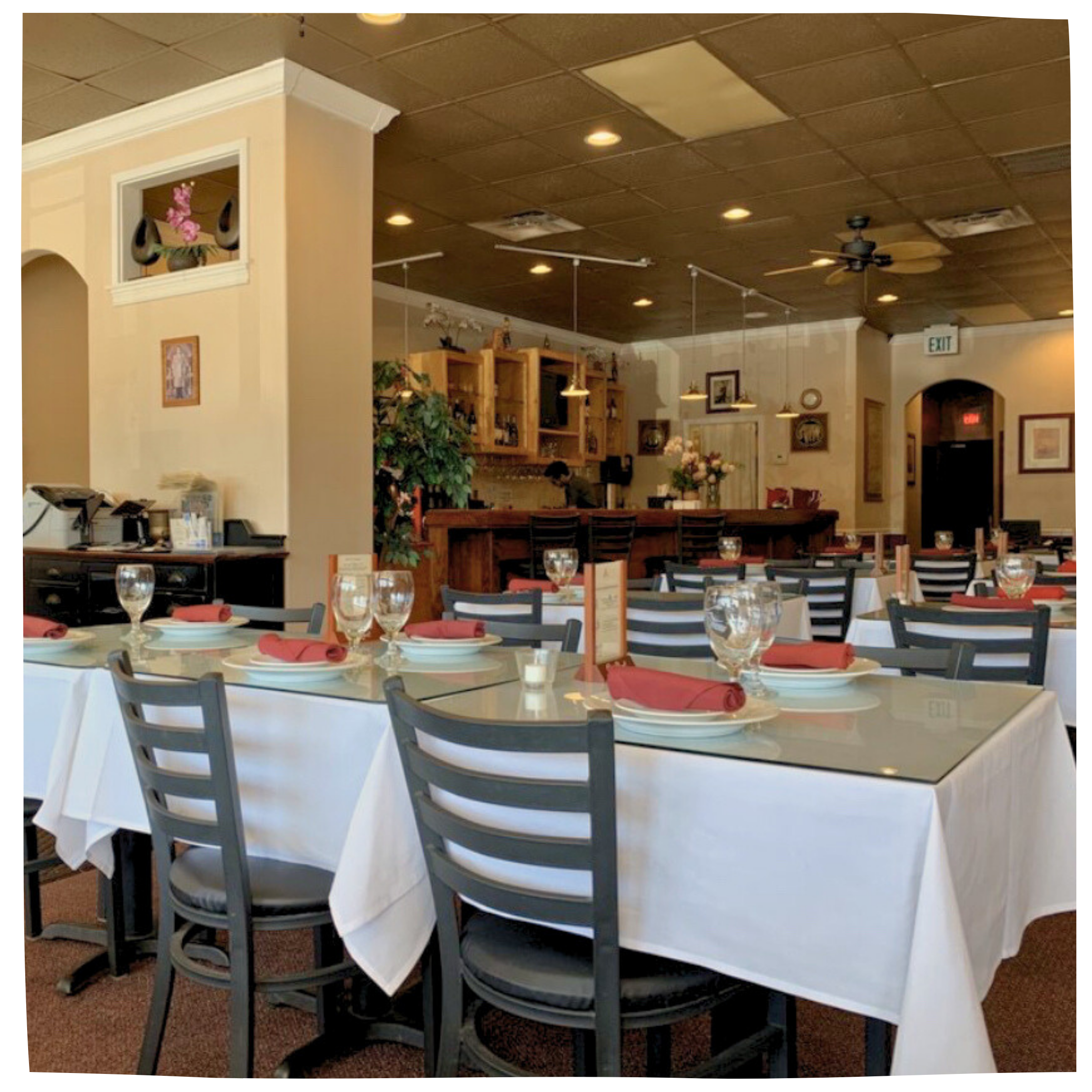 Interior of a restaurant with tables set with white tablecloths, red napkins, glasses, and plates, featuring a bar area in the background and warm lighting.