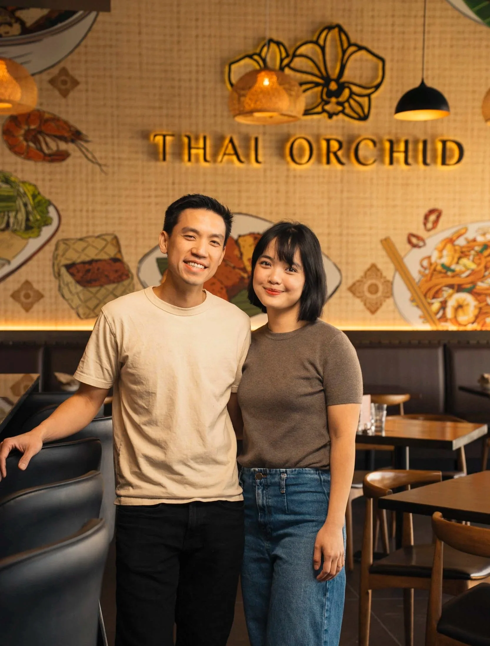 Two people standing inside a Thai restaurant named 'Thal Orchid', with a decorated wall featuring orchids, food illustrations, and warm lighting.