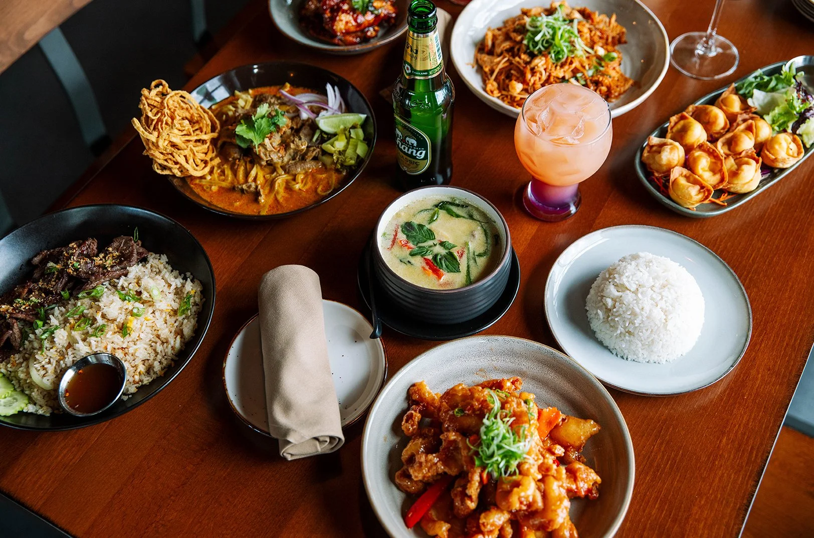 A variety of Asian dishes on a wooden table, including rice, stir-fried chicken, curry, noodle soup, dumplings, and drinks.