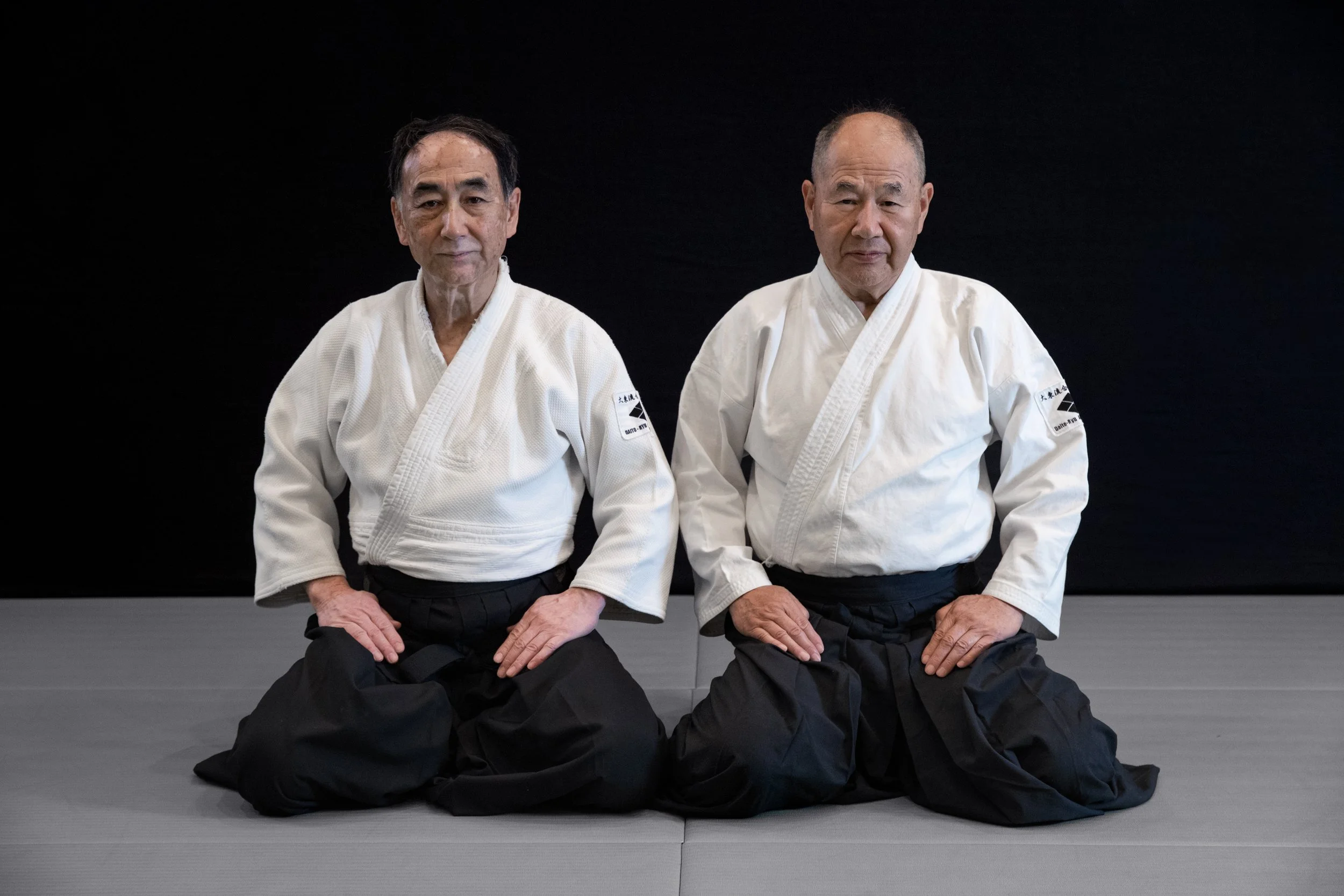 Two elderly men in traditional white martial arts uniforms practicing judo on a gray mat against a black background.