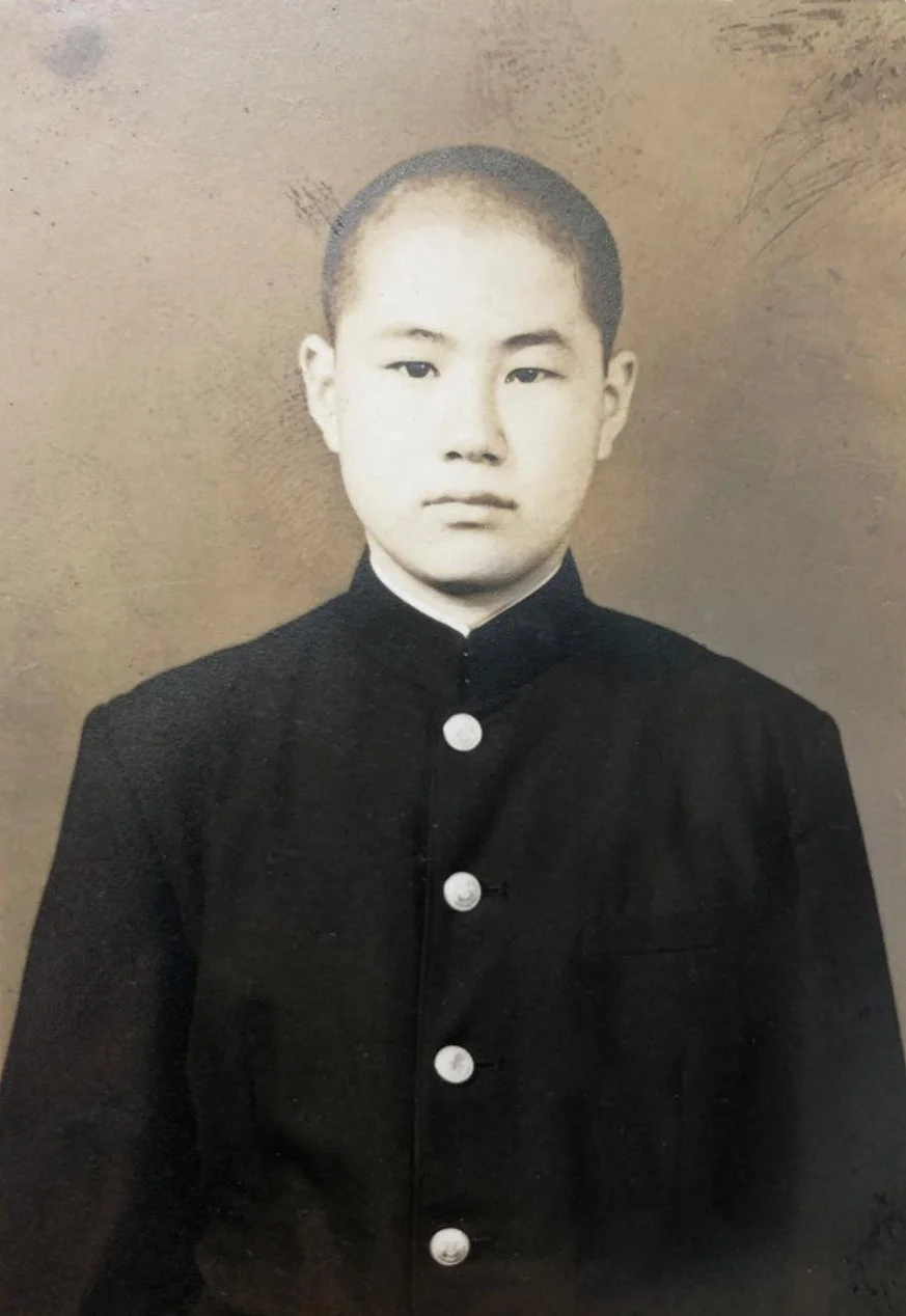 A black-and-white portrait of a young Asian boy wearing a traditional Japanese school uniform with a high collar and white buttons, standing against a plain background.