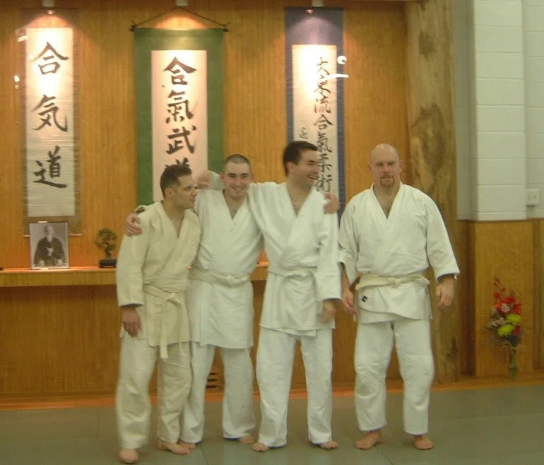 Four men in judo uniforms standing together in a dojo, with Japanese banners on the wall behind them.