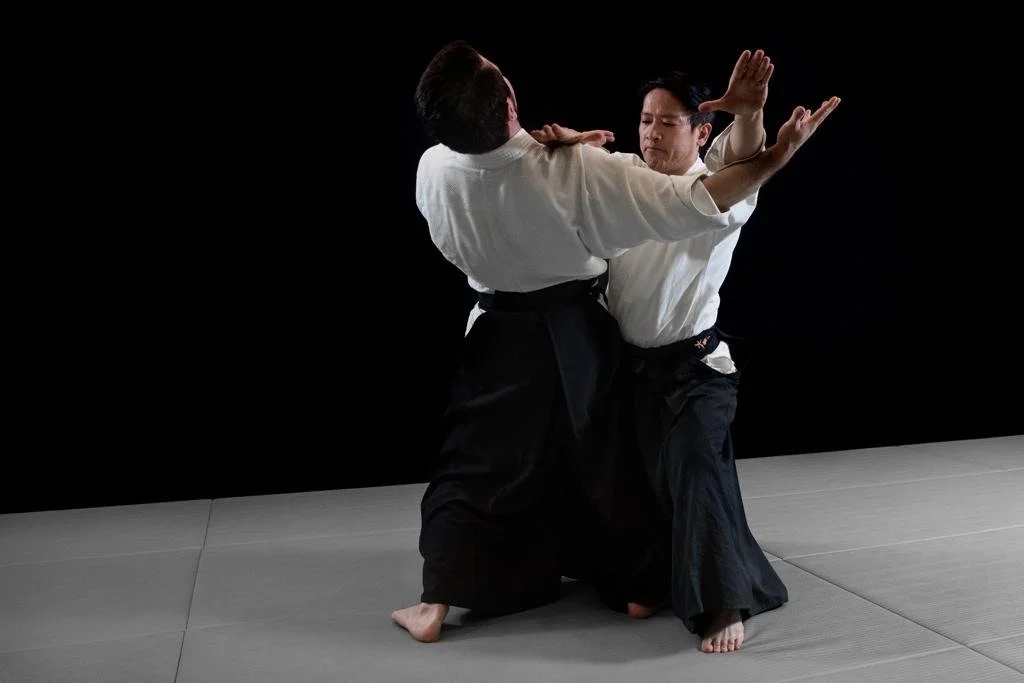 Two martial artists practicing Aikido on a gray mat, one executing a technique on the other, against a black background.