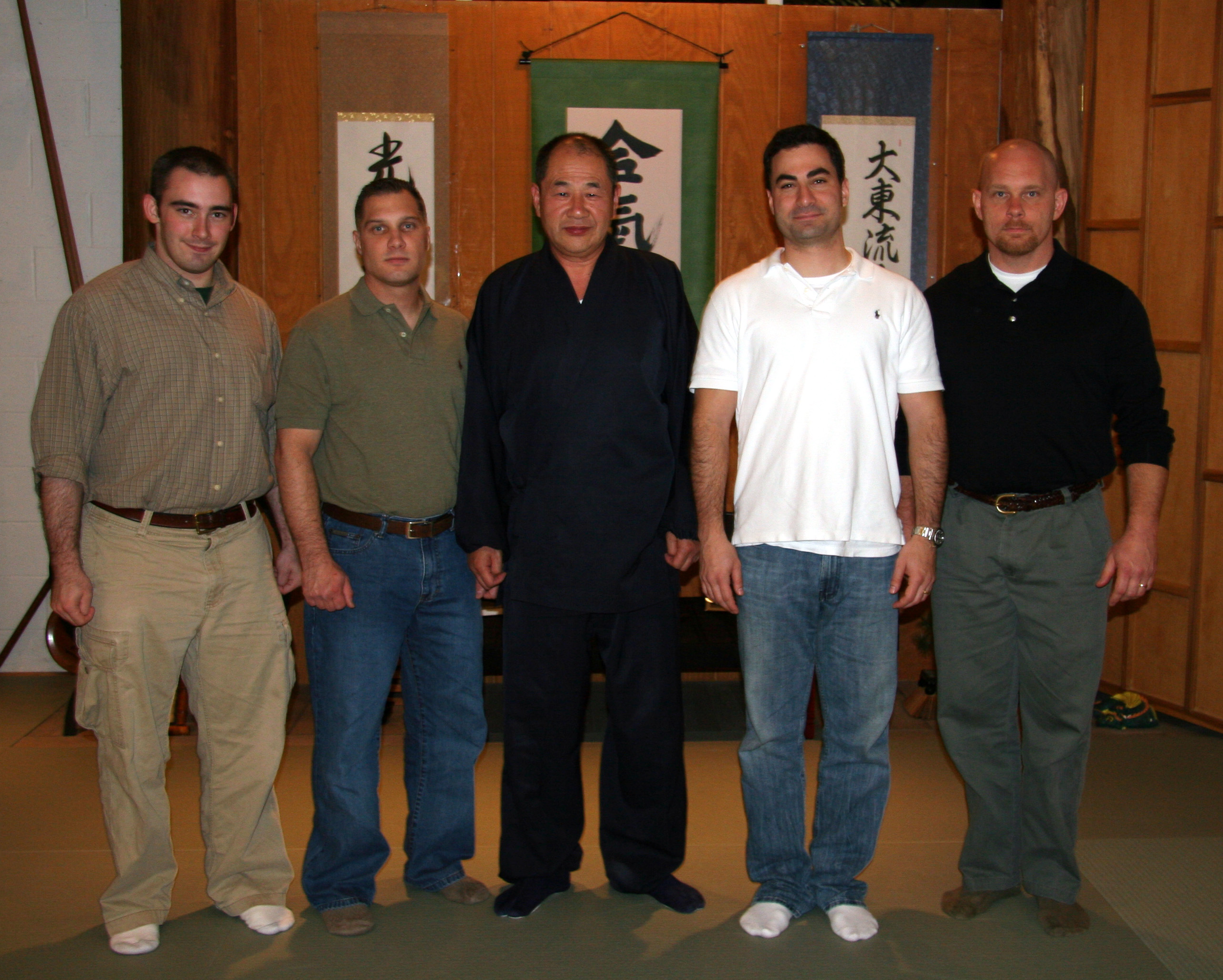 Five men standing side by side indoors with Japanese calligraphy banners in the background.