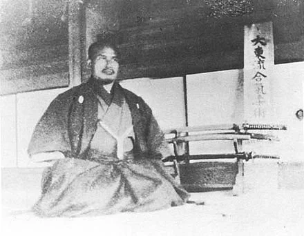Black and white photo of a woman in traditional Japanese clothing sitting on the floor, with swords displayed behind her, and a banner with Japanese characters hanging on the wall.