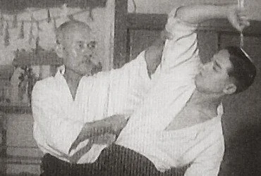 An elderly man demonstrating or practicing martial arts or self-defense with a younger man, indoors.