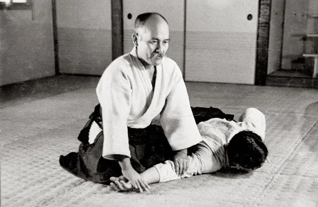A man in traditional Japanese martial arts attire practicing a technique on a woman lying on the floor, in a dojo setting.