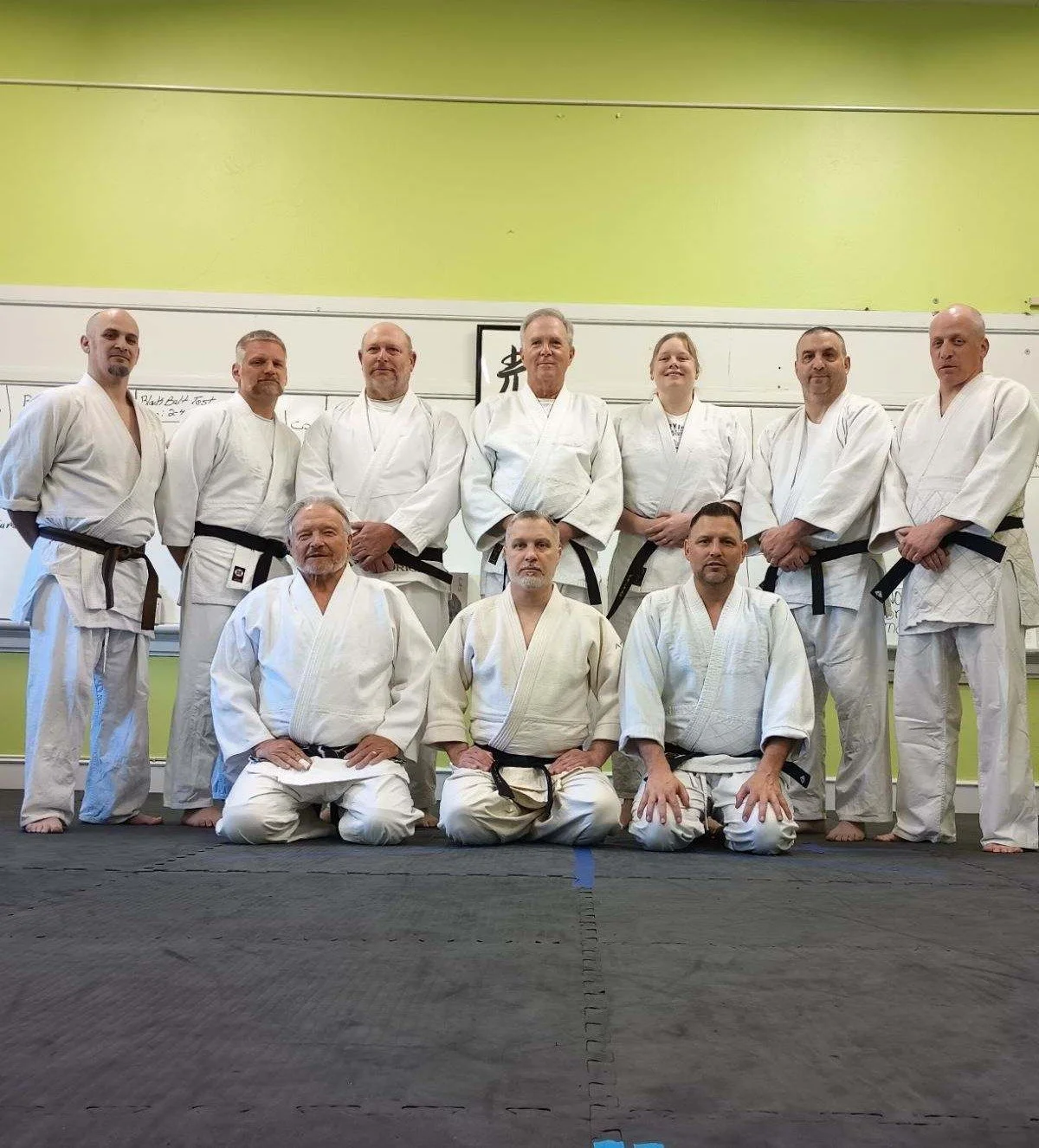 Group of people in martial arts uniforms (gis) posing for a photo in a dojo, some standing and some kneeling, on a black mat.