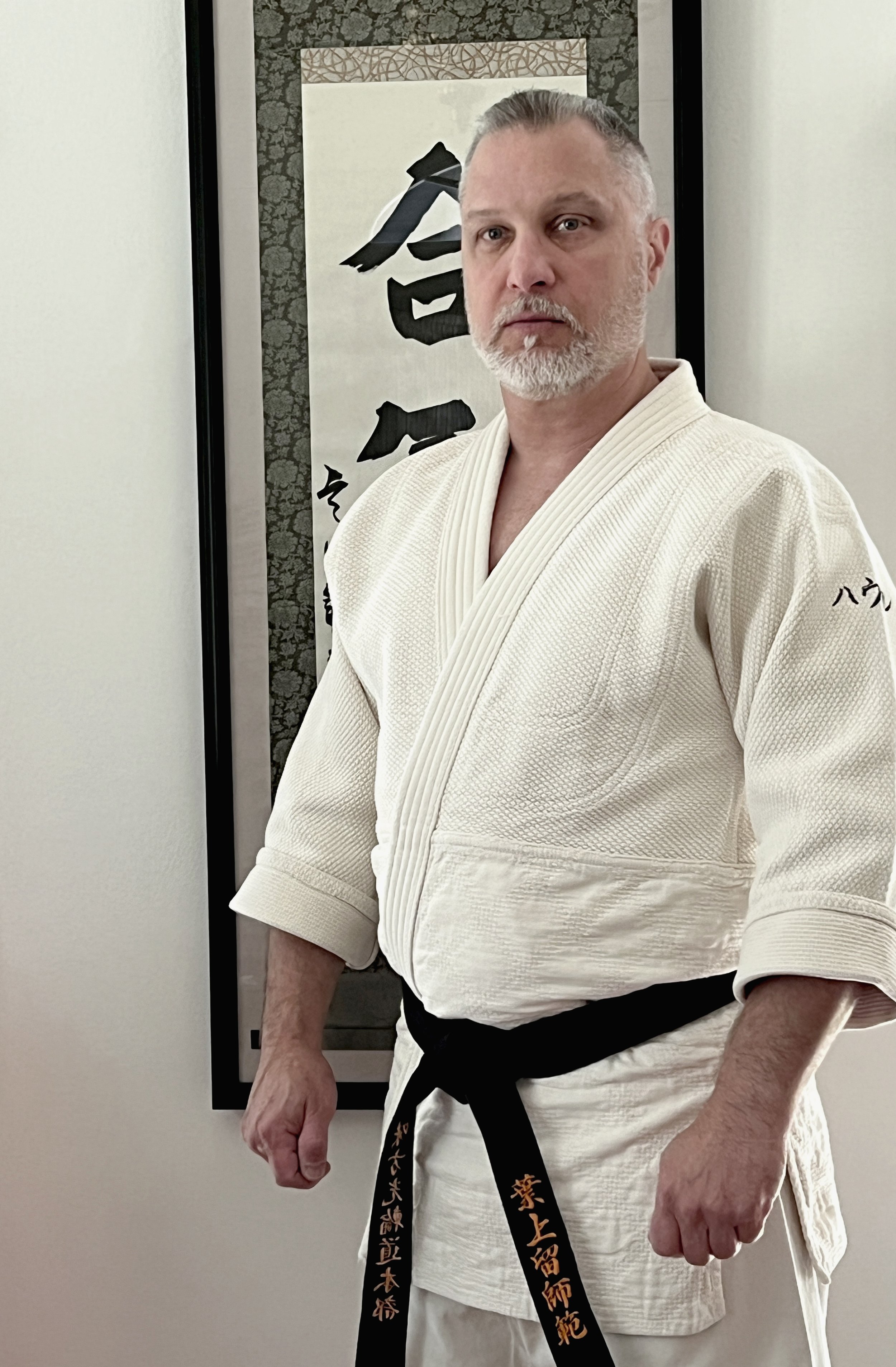 A man wearing a white martial arts gi with a black belt, standing in front of a piece of calligraphy art on the wall.