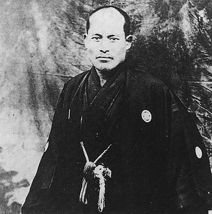 A black and white portrait of a man dressed in traditional Japanese clothing, standing outdoors with trees in the background.