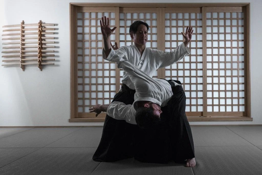 Two martial artists practicing Aikido in a dojo with a wooden lattice window, one in a white gi executing a move on the other in a black hakama.
