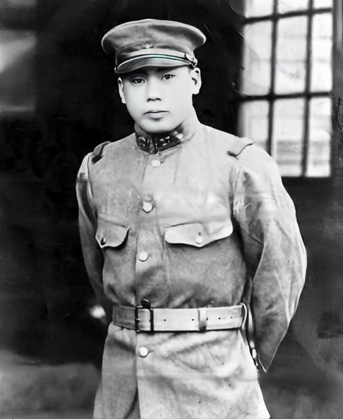 Black and white photo of a young man in a military uniform and cap, standing indoors near a window.
