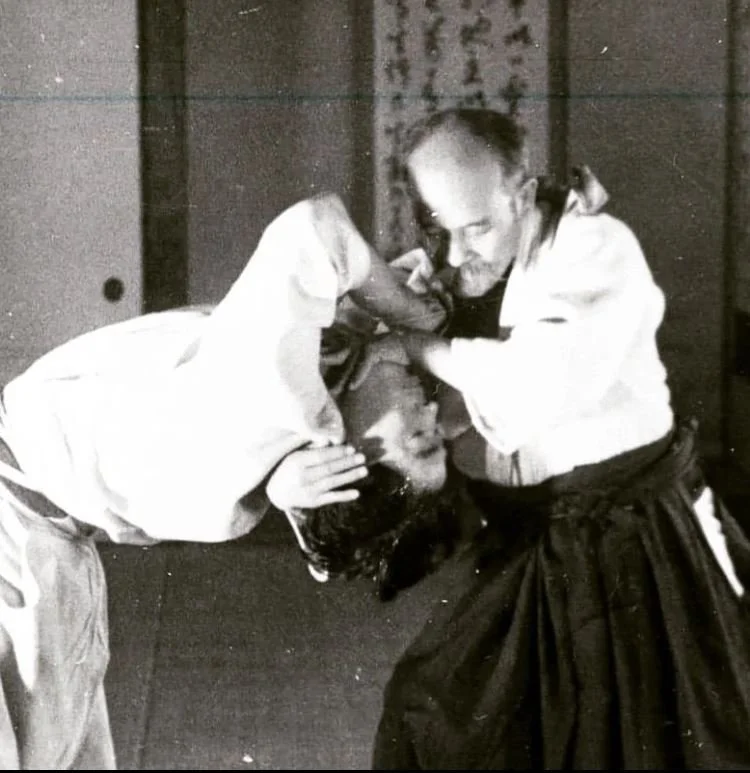 Two martial artists practicing Aikido, wearing traditional gi uniforms, one with a black hakama, in a dojo setting.