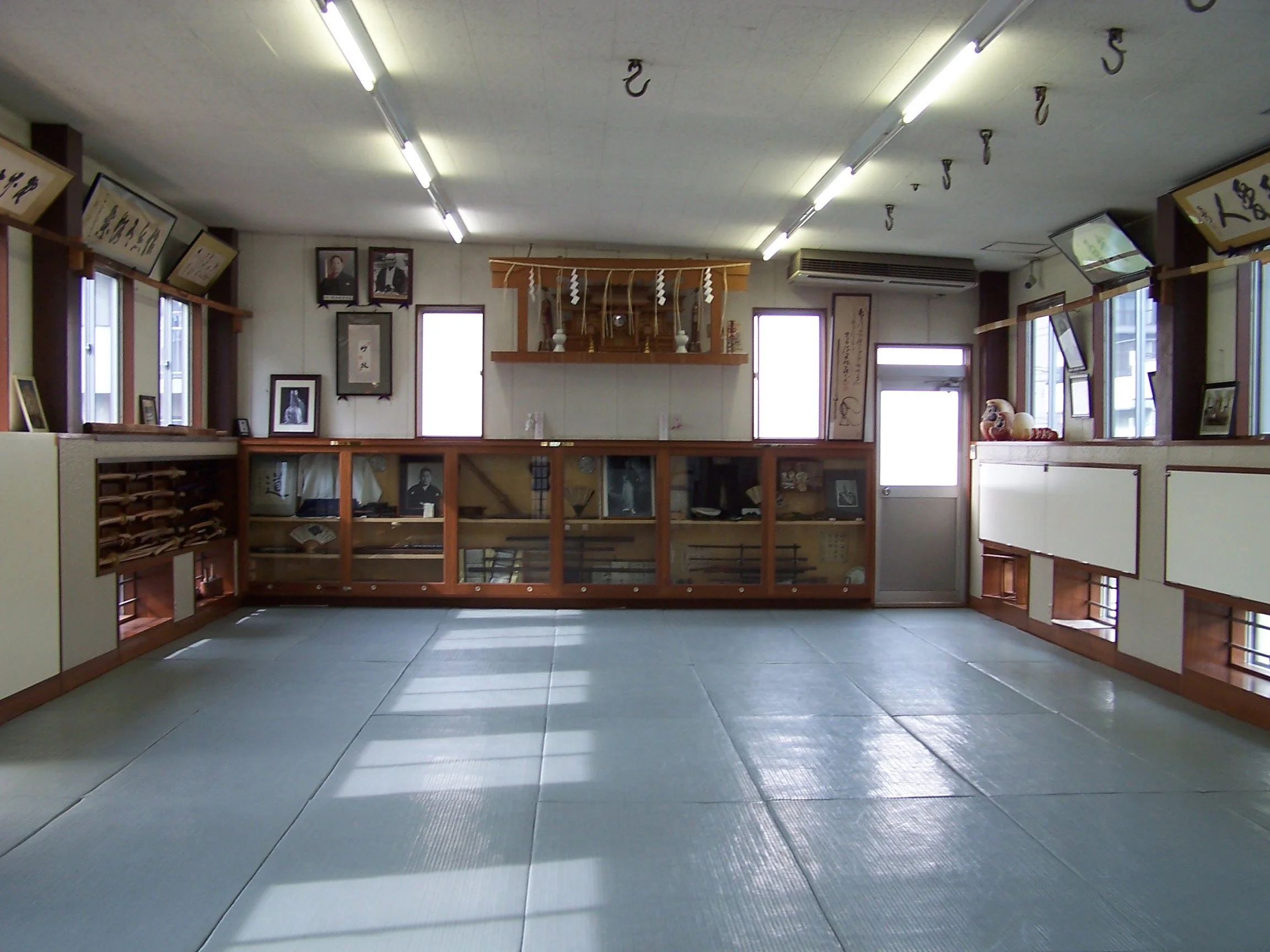 Interior of a traditional Japanese dojo with tatami mats on the floor, wooden display shelves, framed artwork and photographs on the walls, and a small shrine or altar at the back.