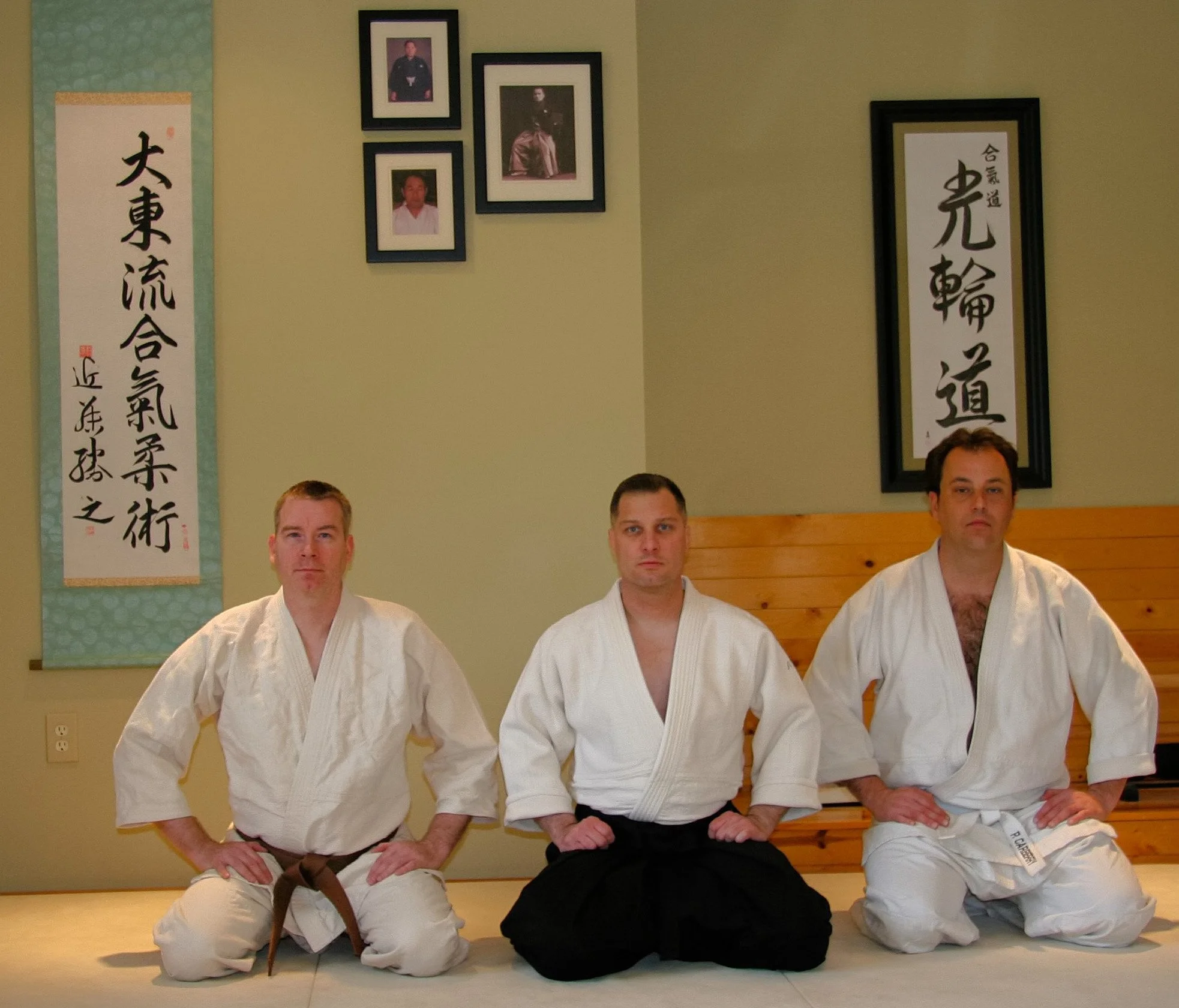 Four martial artists dressed in white gis sitting and kneeling in a dojo, with framed pictures and Japanese calligraphy on the wall behind them.