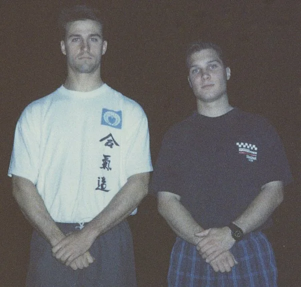 Two young men standing side by side against a dark background, wearing casual T-shirts and looking at the camera.