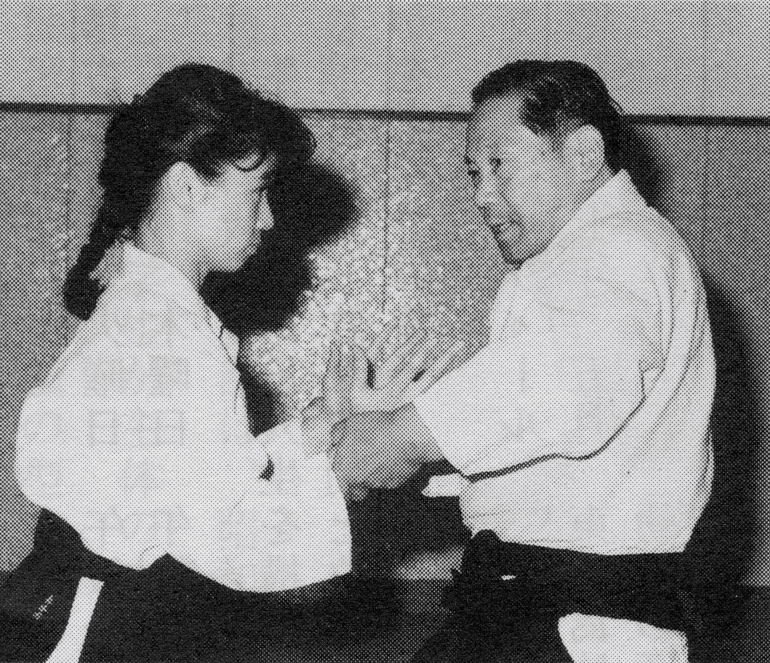 A black and white halftone image depicting a young girl with long hair and a young boy with glasses, both sitting at a table with a plate of food, smiling and facing each other.