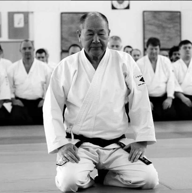 An elderly Japanese man in a martial arts uniform kneeling on a mat, surrounded by students also in martial arts uniforms, in a dojo.