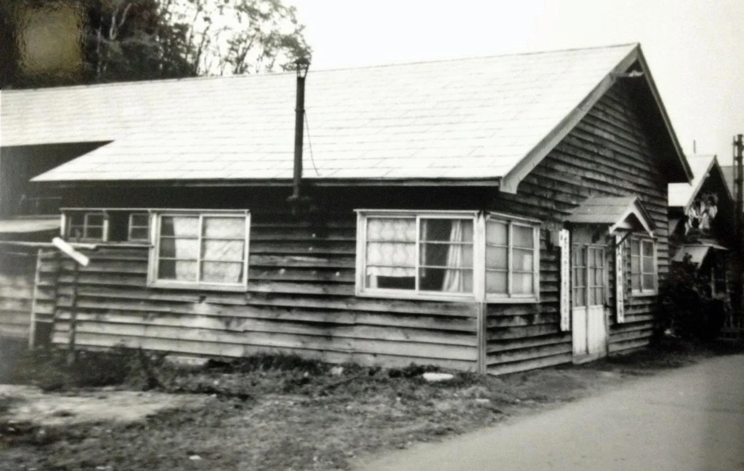 Black and white photo of a wooden house with a sloped roof and multiple windows, surrounded by unpaved ground and some trees in the background.