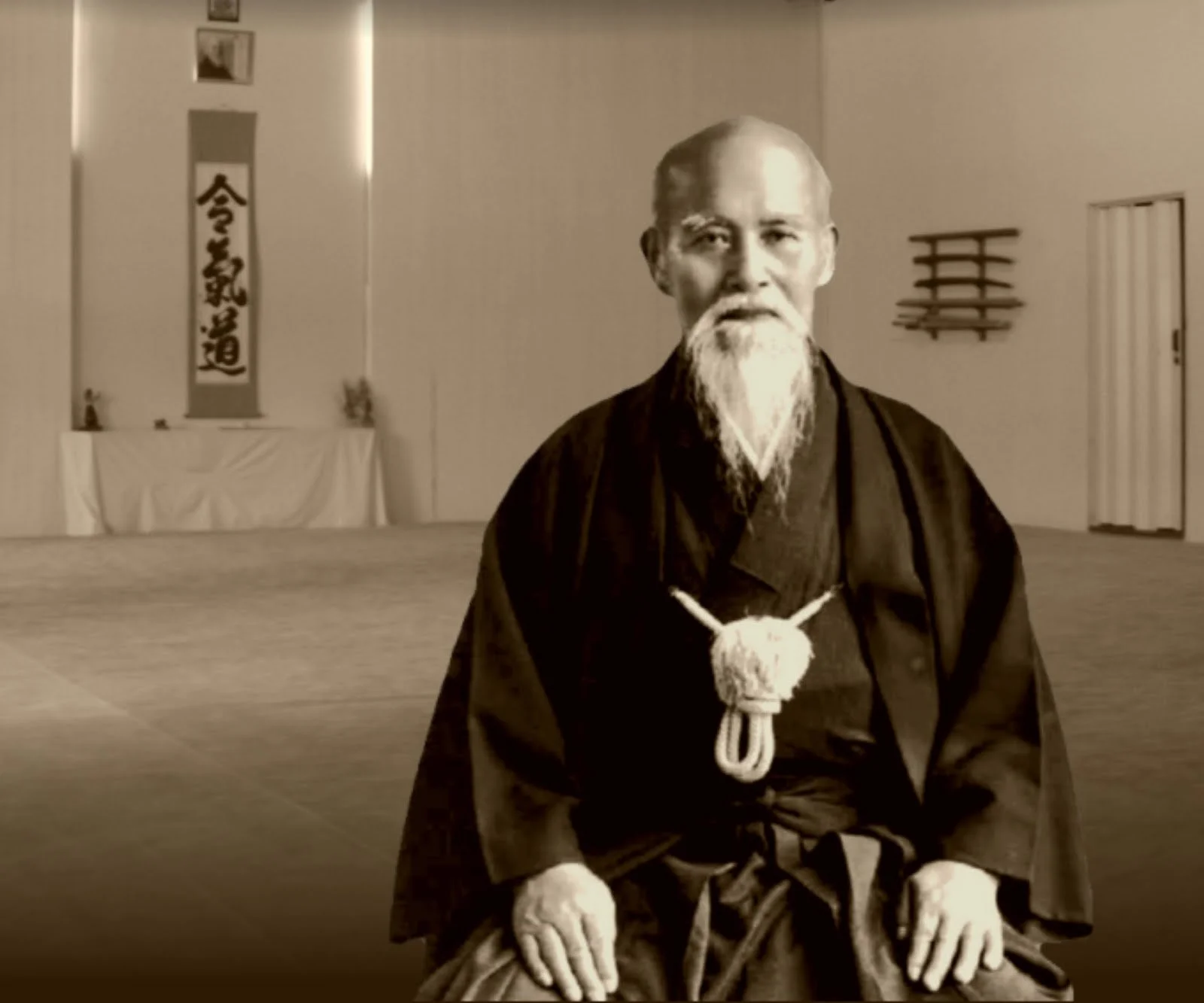 A sepia-toned photograph of an elderly Asian man with a long white beard, dressed in traditional Japanese martial arts clothing, sitting in a dojo with katanas on the wall and a Japanese scroll in the background.