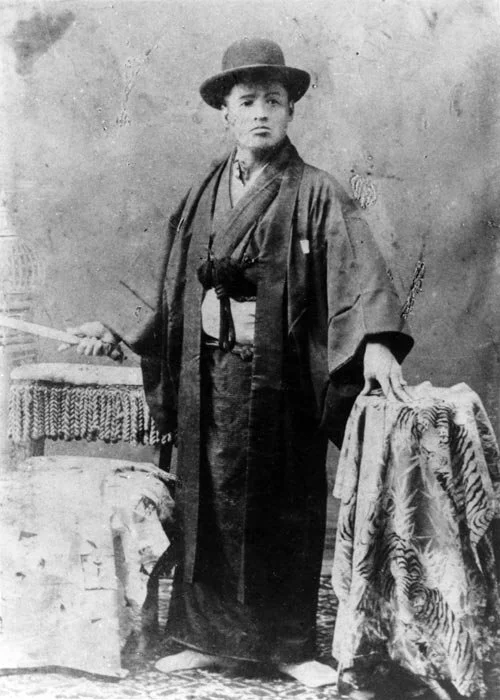 Black and white photo of a young boy in traditional Japanese clothing and hat, standing between two tables with ornate tablecloths, holding objects in each hand.