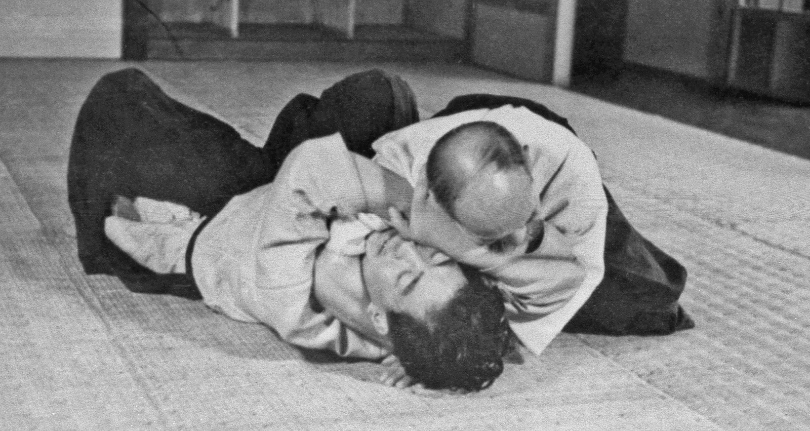 Two martial artists in gi uniforms practicing a grappling technique on a tatami mat.
