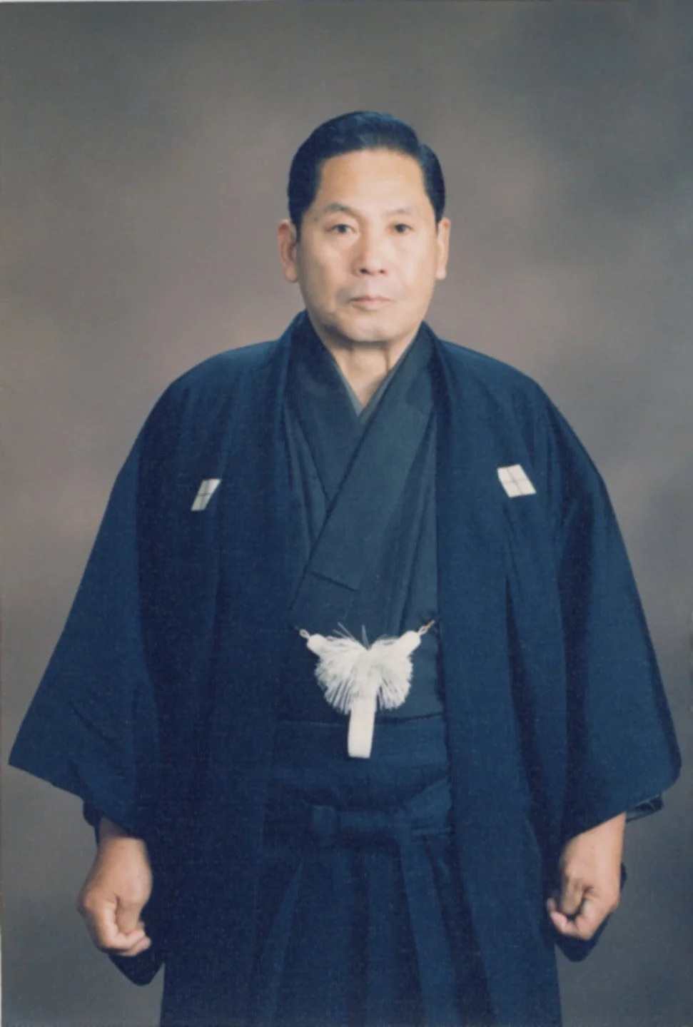 A man in traditional Japanese attire, wearing a black kimono with a white tassel accessory, standing against a plain background.