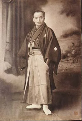 A black and white photograph of a man dressed in traditional Japanese clothing, standing outdoors with a landscape background.