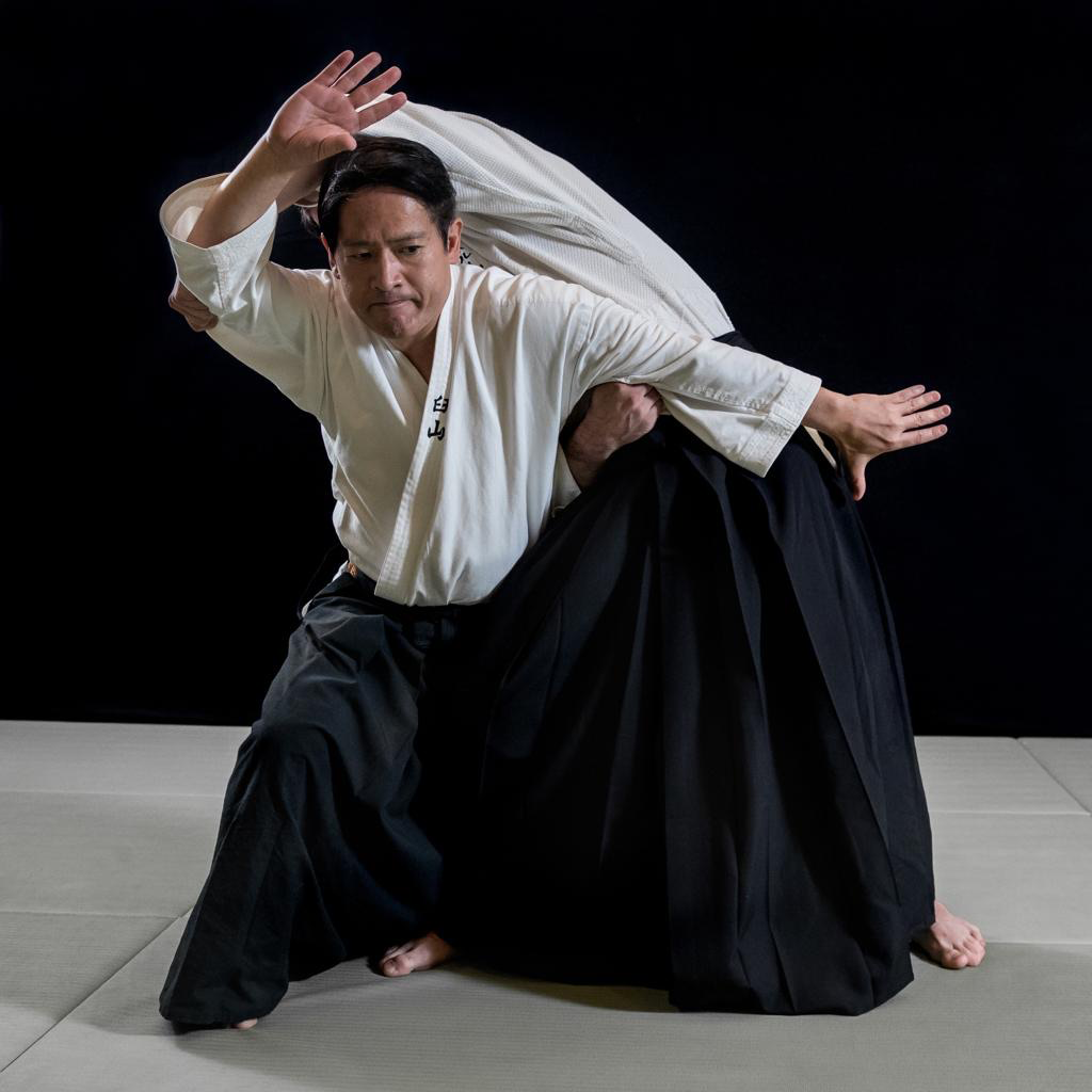 Two martial artists practicing a judo throw, one dressed in a white gi and the other in a black gi, against a black background.