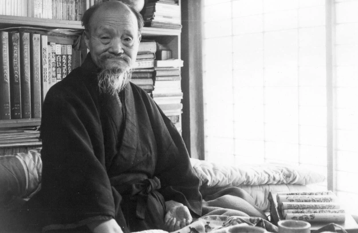 An elderly man with a beard and mustache, wearing traditional Japanese clothing, sitting in a room with bookshelves and stacked books, smiling at the camera.
