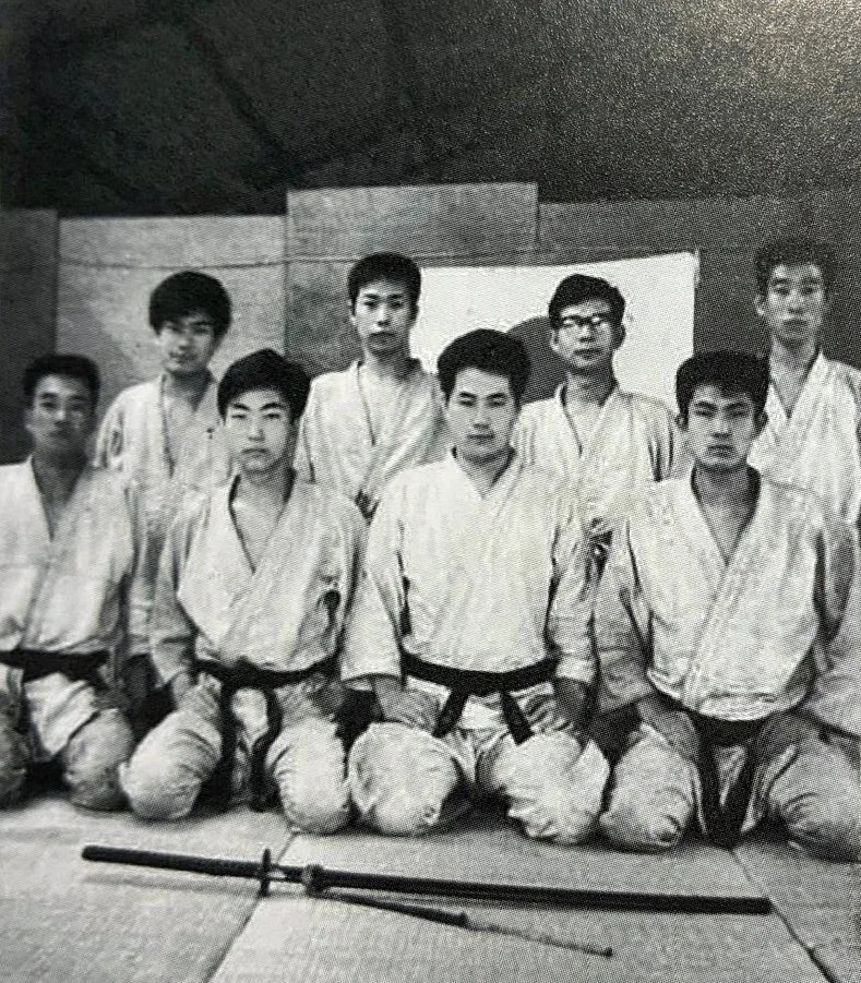 Group of martial artists in gi uniforms posing in a dojo with weapons in front of them.