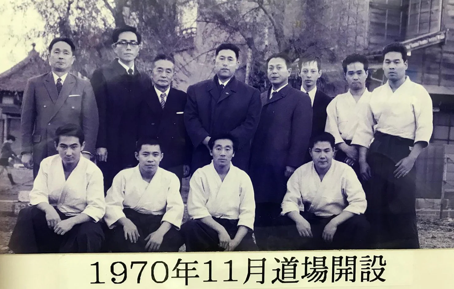 Black and white group photo of nine men, some in suits and some in martial arts uniforms, outdoors with trees and traditional buildings in the background. The photo is dated November 1970.