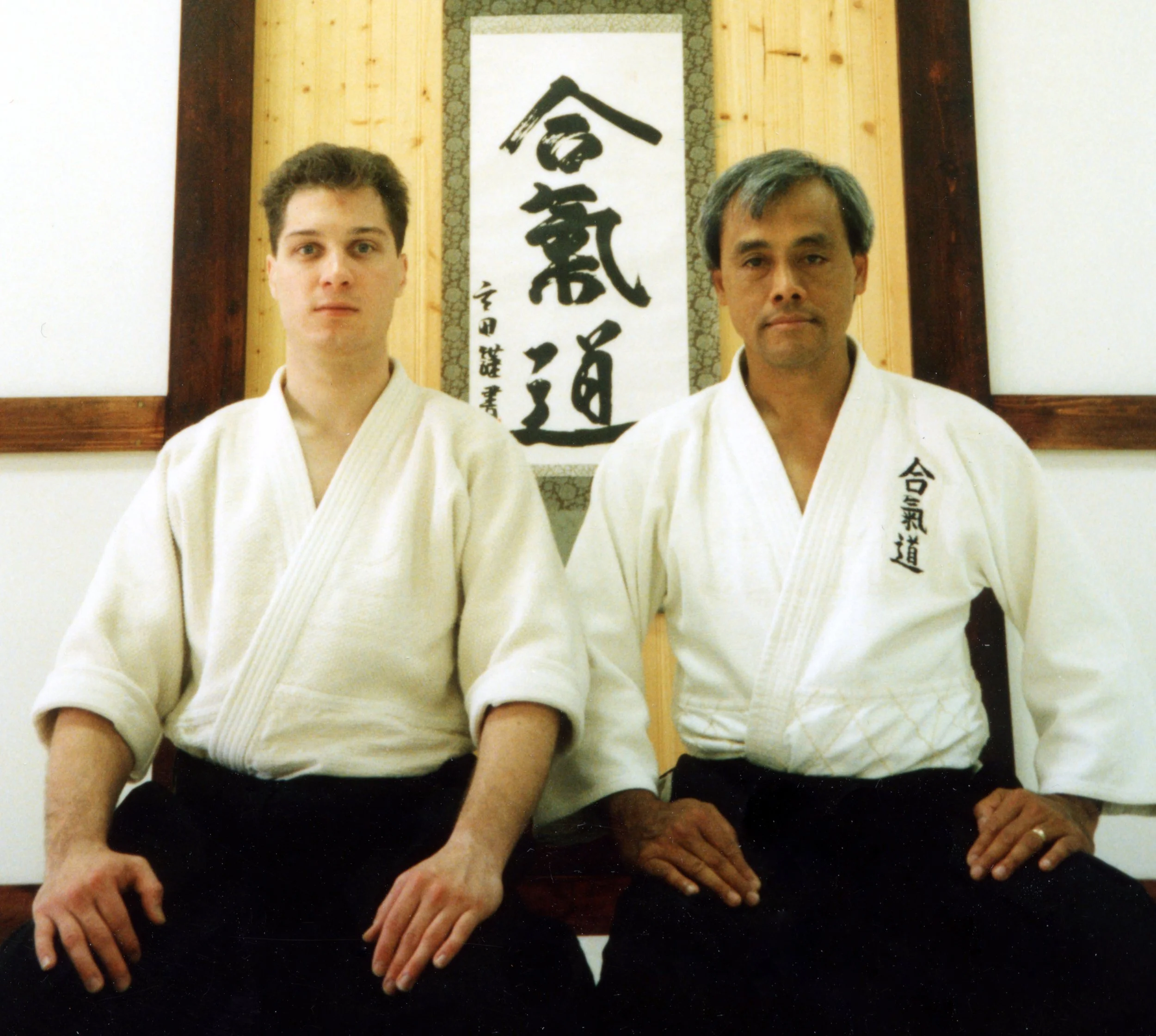 Two men wearing white martial arts uniforms standing side by side in front of Japanese calligraphy art. The man on the left is younger with short, dark hair, and the man on the right appears older with gray hair.