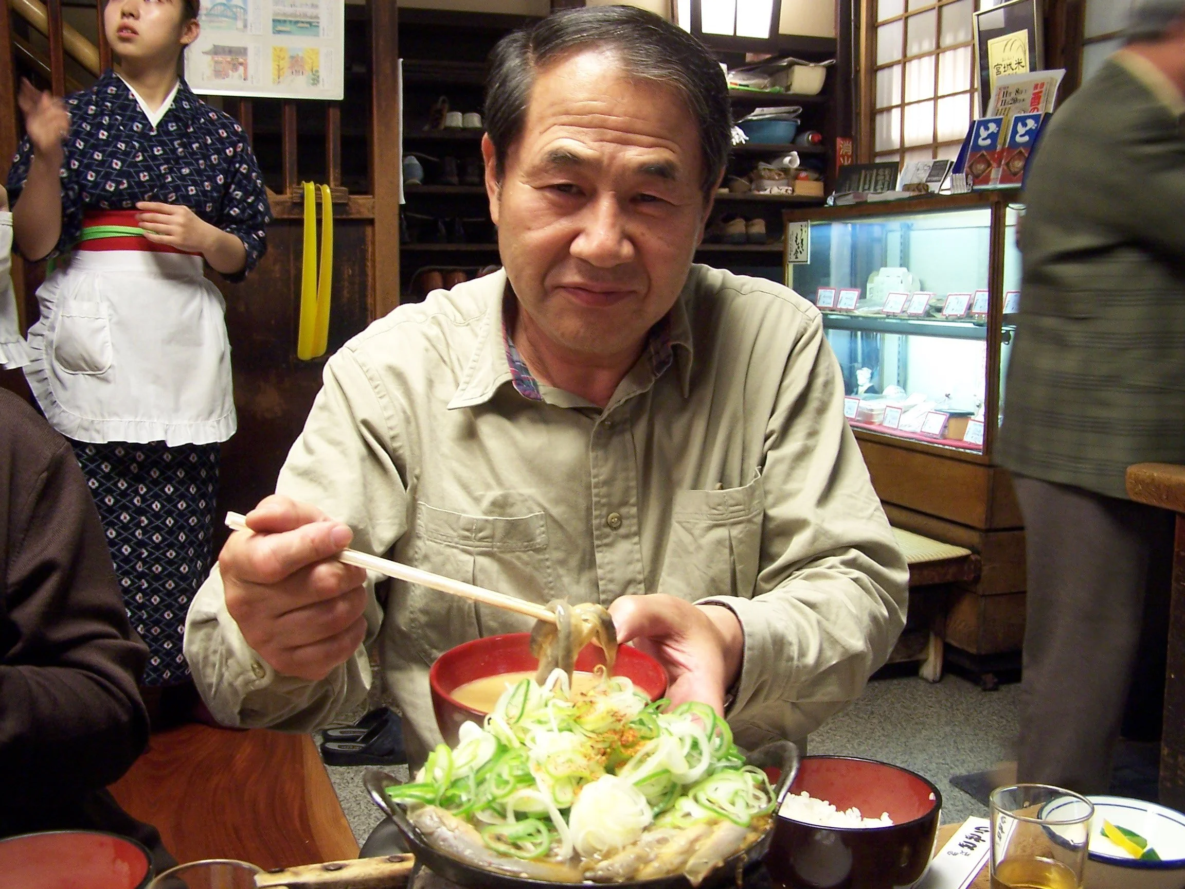 A middle-aged man eating from a bowl of noodles using chopsticks in a Japanese restaurant, with a woman in traditional yukata standing nearby and a display of shoes on shelves in the background.