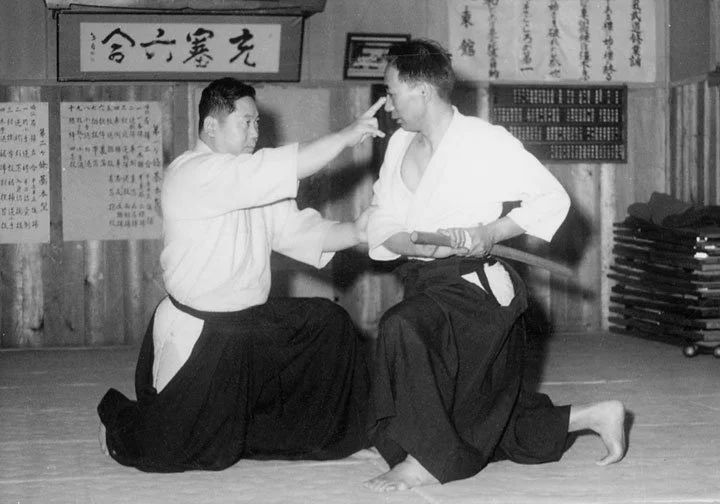 Two martial artists practicing in a dojo, one is adjusting the other's face while the other holds a training sword, both wearing traditional uniforms.