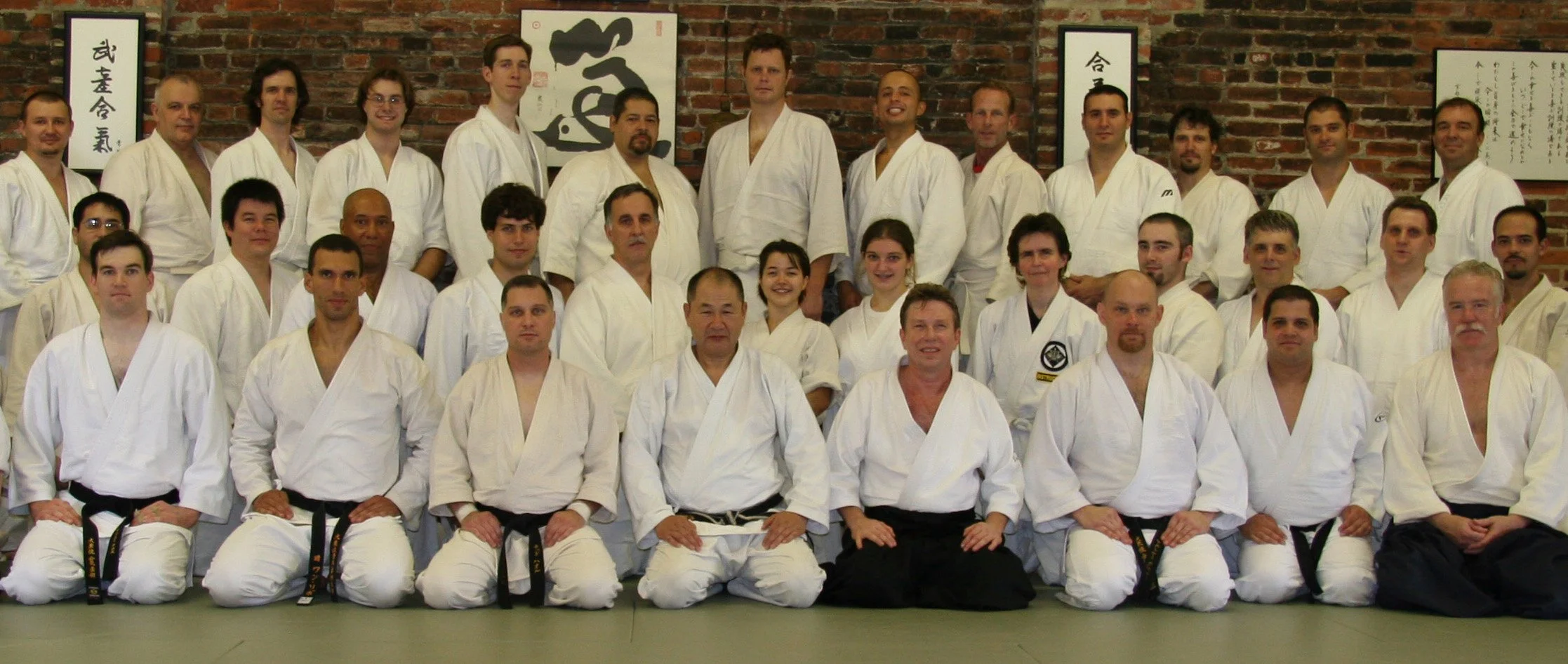 Group photo of martial arts practitioners in white gis, kneeling and standing in front of a brick wall adorned with Japanese calligraphy.