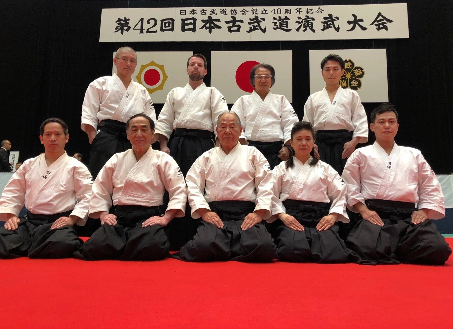 Group of martial arts practitioners in white gis with black belts posing for a photo on a red mat, with a banner and flags in the background commemorating the 40th anniversary of a Japanese martial arts association.