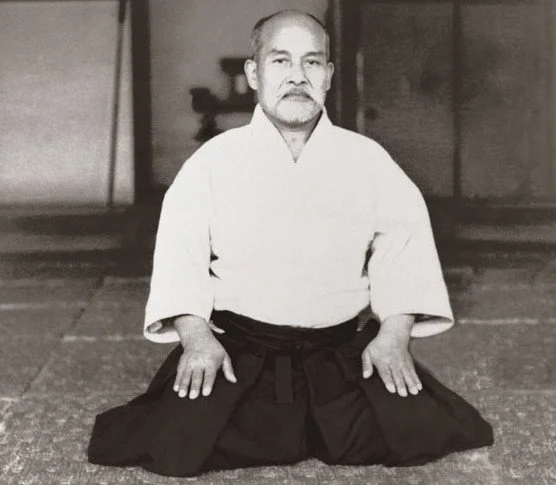 A man in traditional martial arts attire kneeling on the floor, indoors, in a dojo or training hall.