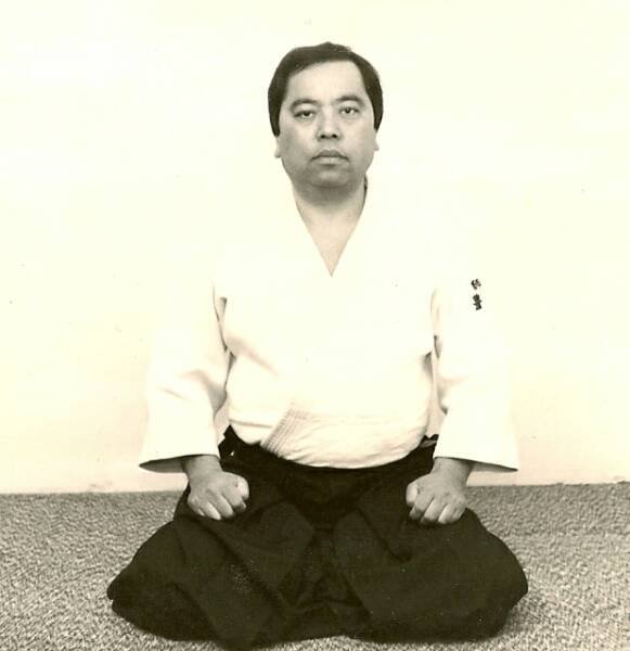 A man in traditional martial arts attire sitting in seiza position against a plain wall.