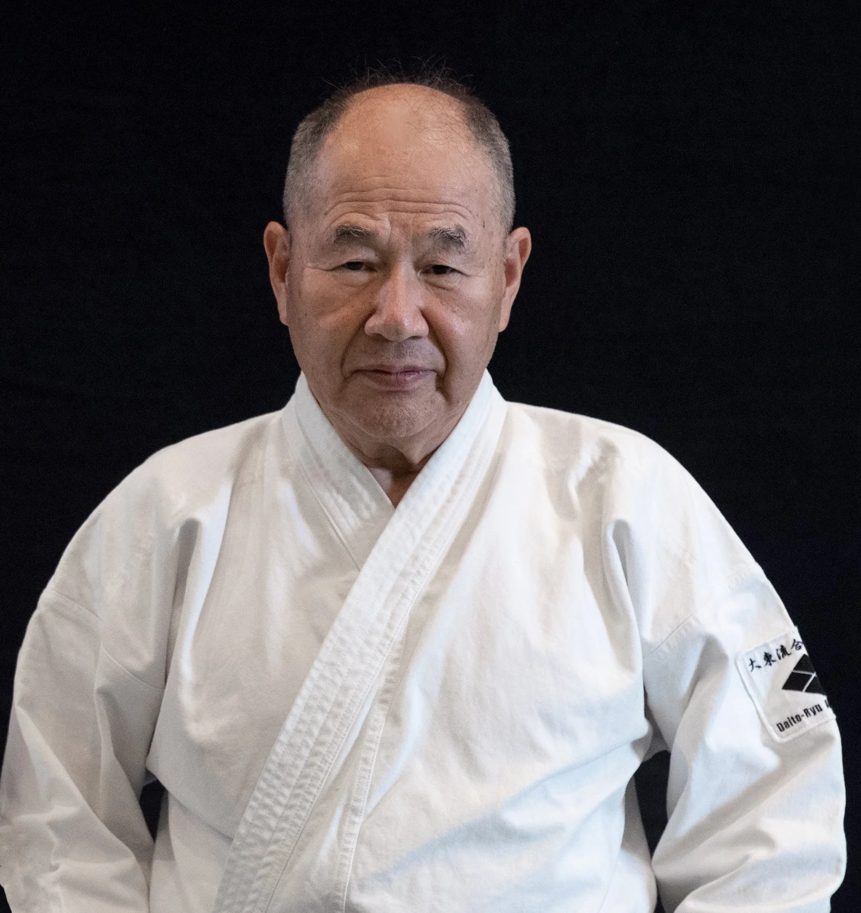 Close-up of an elderly person wearing a white martial arts uniform, sitting in a meditative pose, against a plain black background.
