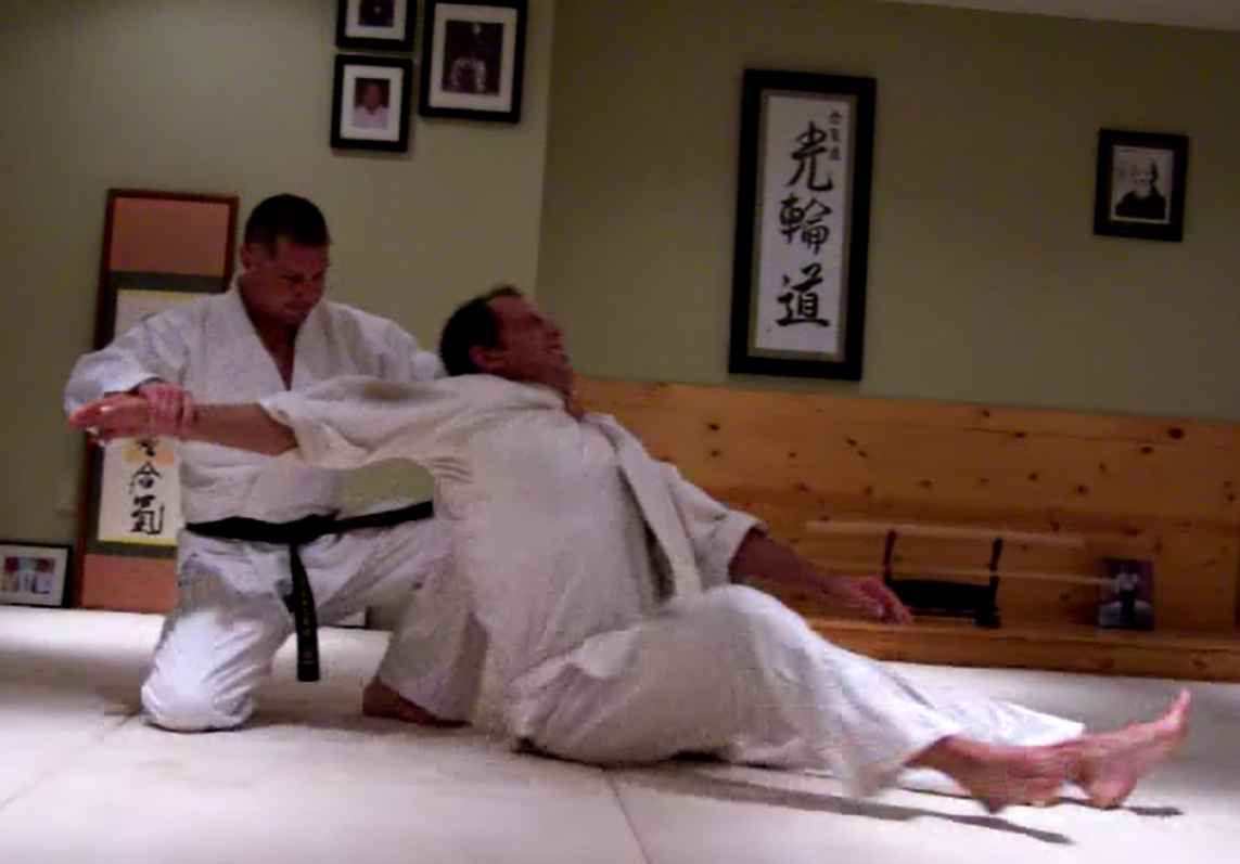 Two martial artists practicing karate or judo on a mat in a dojo. One is in a dominant position on the floor, while the other is standing and controlling their opponent.