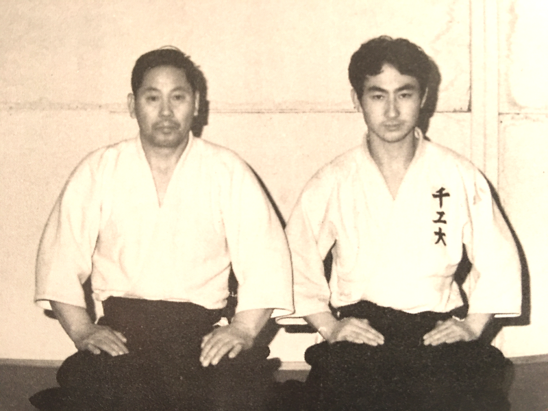 Two young men dressed in traditional martial arts uniforms sitting on a mat against a plain wall, possibly in a dojo.