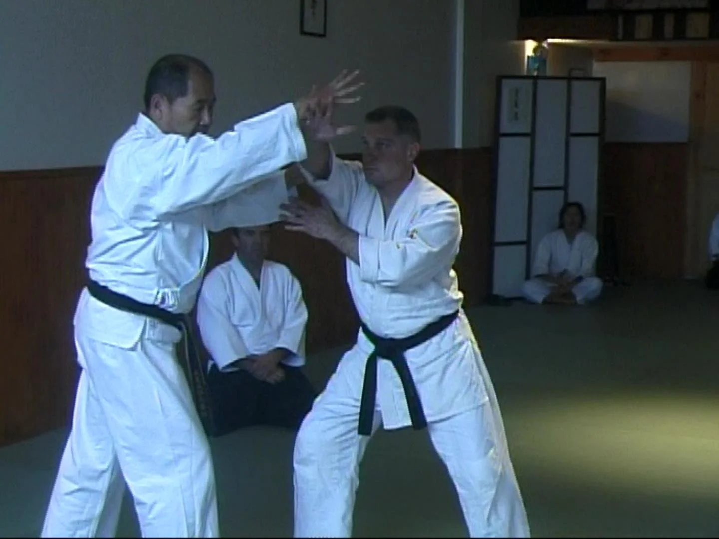 Two martial artists practicing judo in a dojo, with one demonstrating a throw on the other while three people observe, two sitting on the floor and one standing in the background.