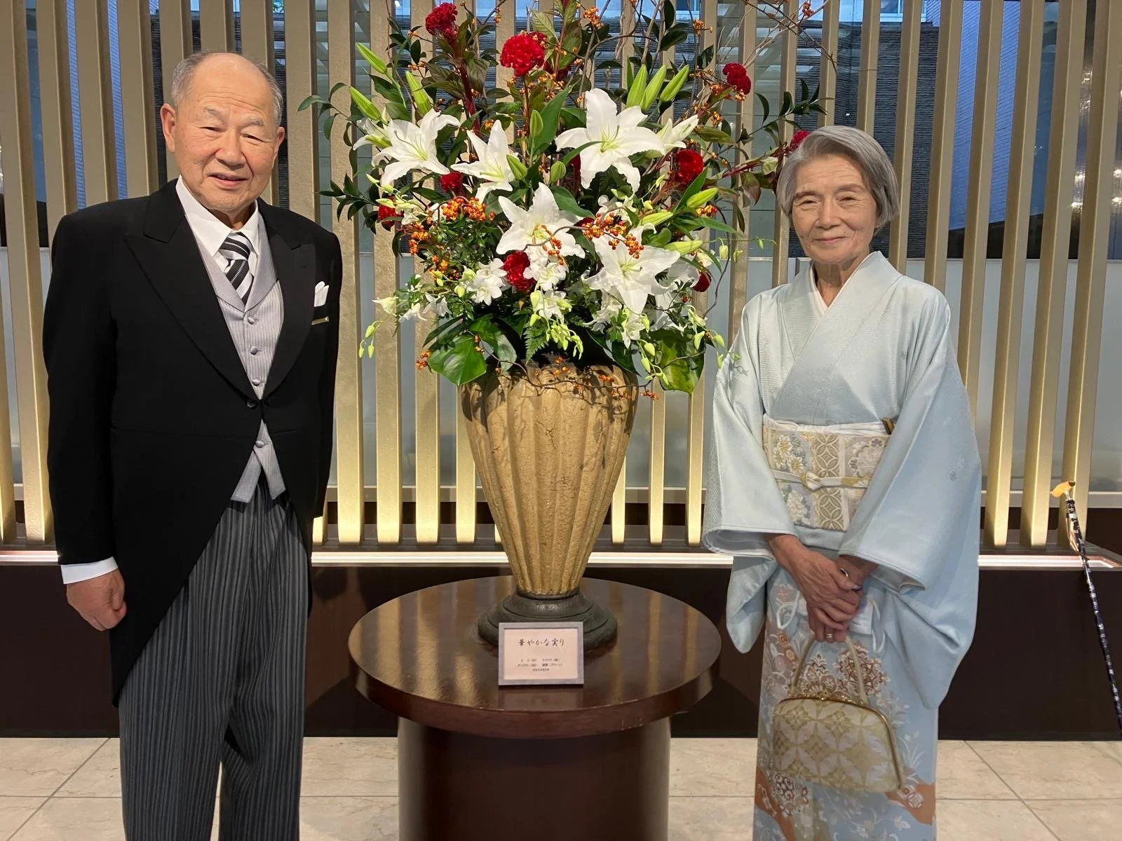 An elderly man in a black tuxedo and elderly woman in a traditional Japanese kimono standing beside a large floral arrangement with white lilies, red flowers, and greenery in a gold vase. They are indoors, in front of a backdrop of vertical gold and silver slats.