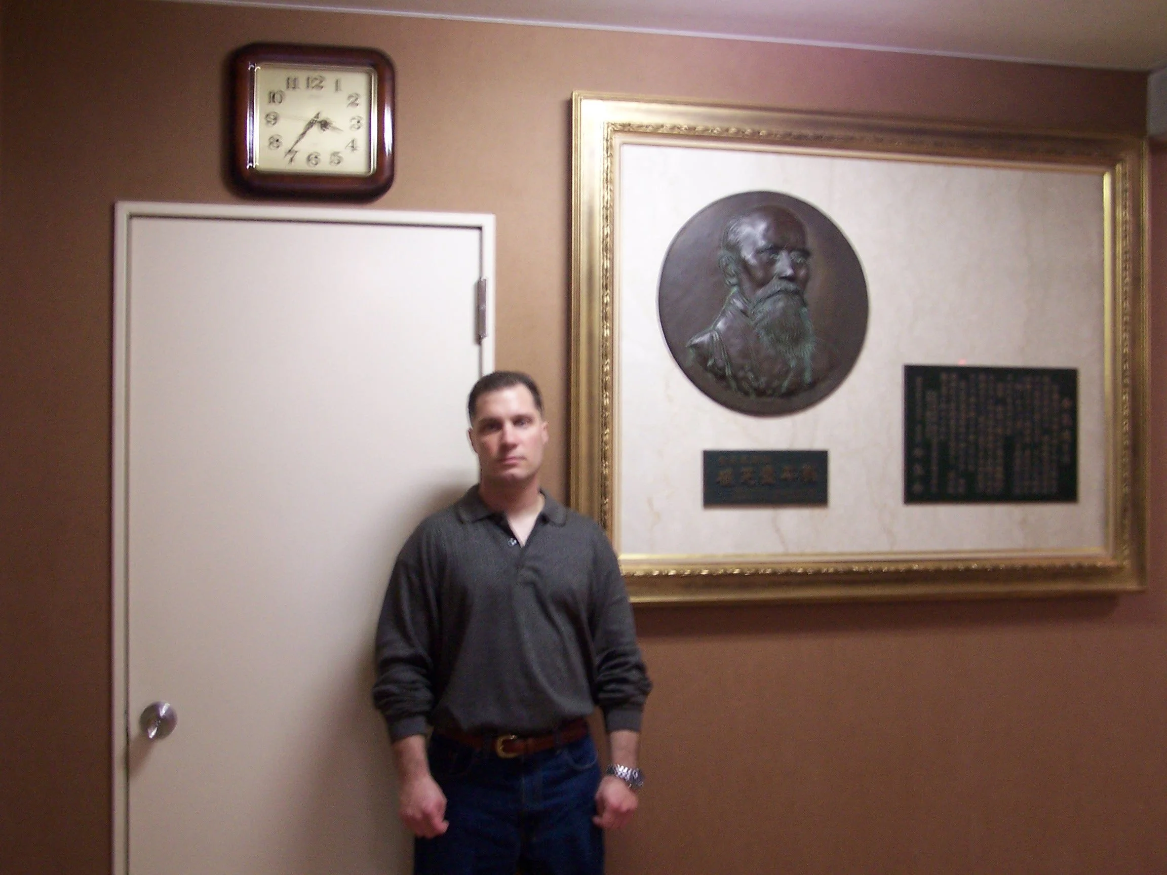 A man with short dark hair and a serious expression, wearing a dark gray shirt, standing in front of a wall with a framed bronze bust of a bearded man, a gold-framed mirror, and plaques, near a white door.